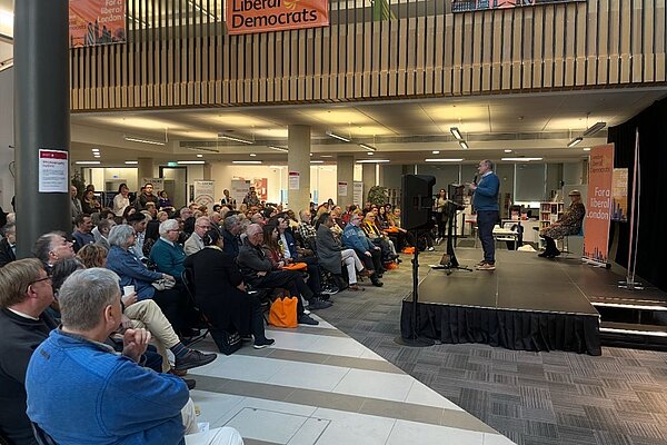 An image of the 2025 London Lib Dem Conference Hall with, on the right hand side, Ed Davey MP speaking, and on the left hand side, an audience of around 80-90 people