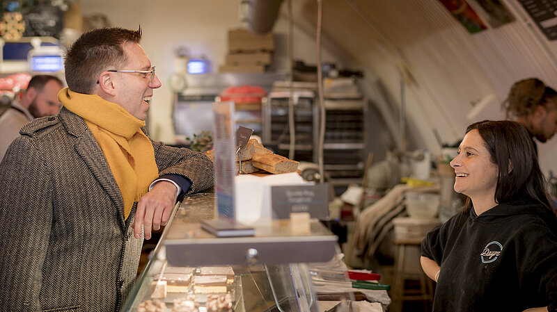 Rob Blackie pictured with a yellow scarf speaking to a bakery owner in Herne Hill, London